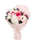 A floral bouquet wrapped in pale pink paper, featuring pink and white roses, white hydrangeas, purple filler flowers, and green foliage, tied with a sheer ribbon at the bottom, on a white background.