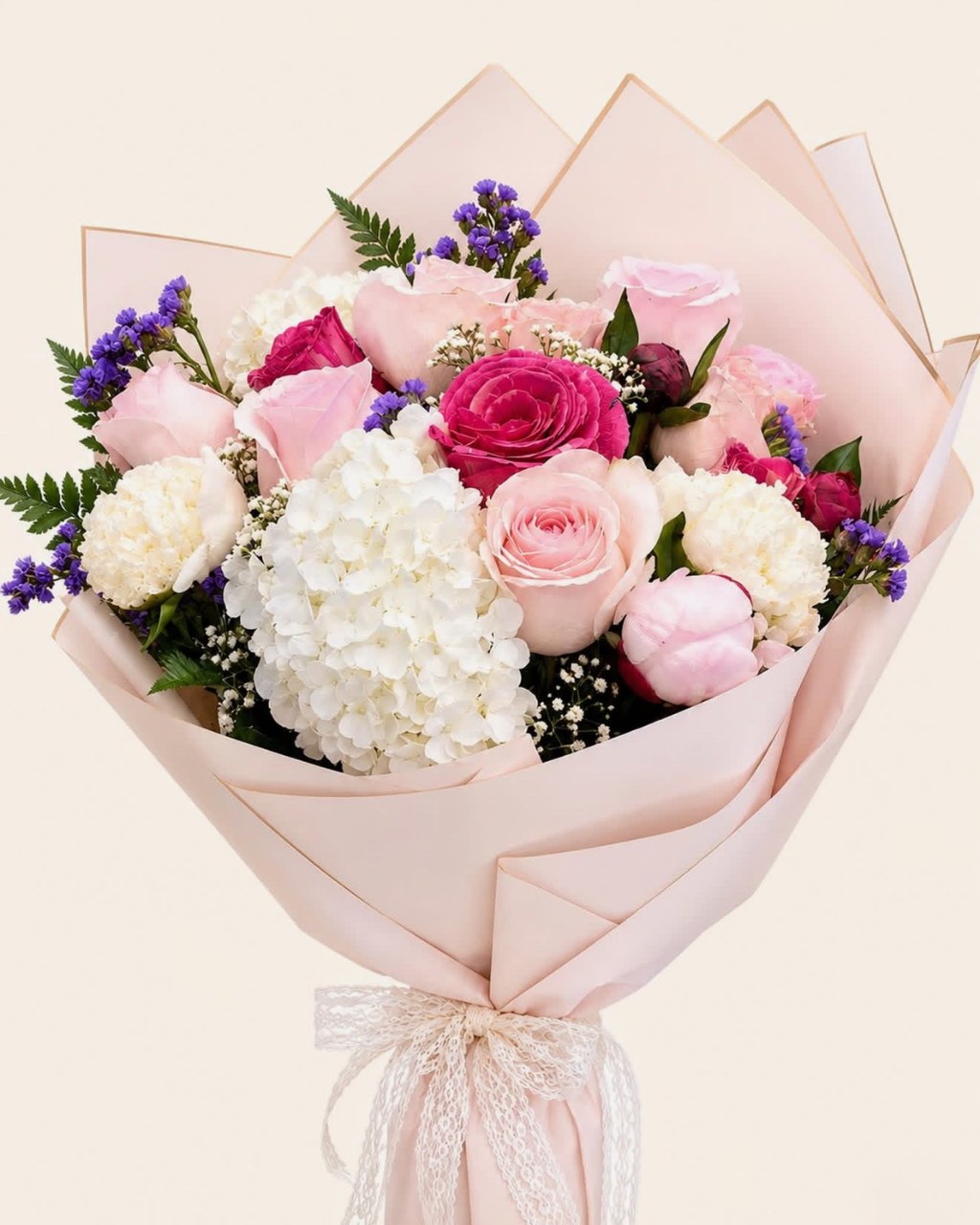 A floral bouquet wrapped in pale blush pink paper, featuring pink and white roses, white hydrangeas, burgundy roses, purple filler flowers, and green foliage, tied with a sheer white ribbon at the bottom.