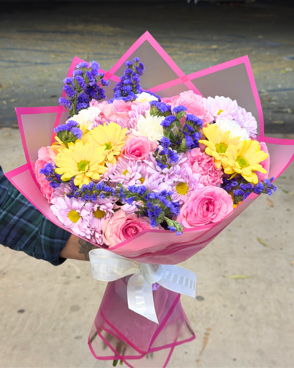 A vibrant flower bouquet wrapped in pink paper with a white ribbon, held by a hand wearing a green plaid shirt. The bouquet features pink roses, yellow daisies, pink and white chrysanthemums, and purple stock flowers, set against an outdoor concrete background.